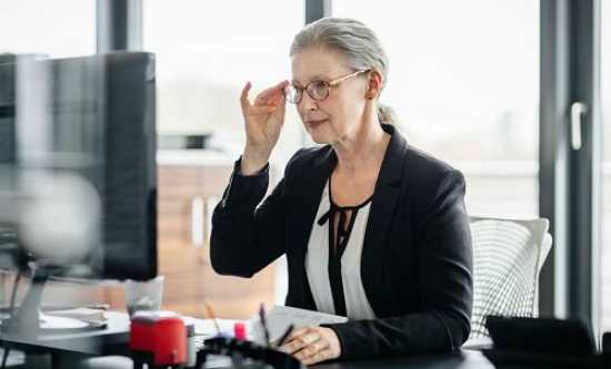 A woman in office clothing looks at her computer monitor, one hand on her glasses as if deep in thought